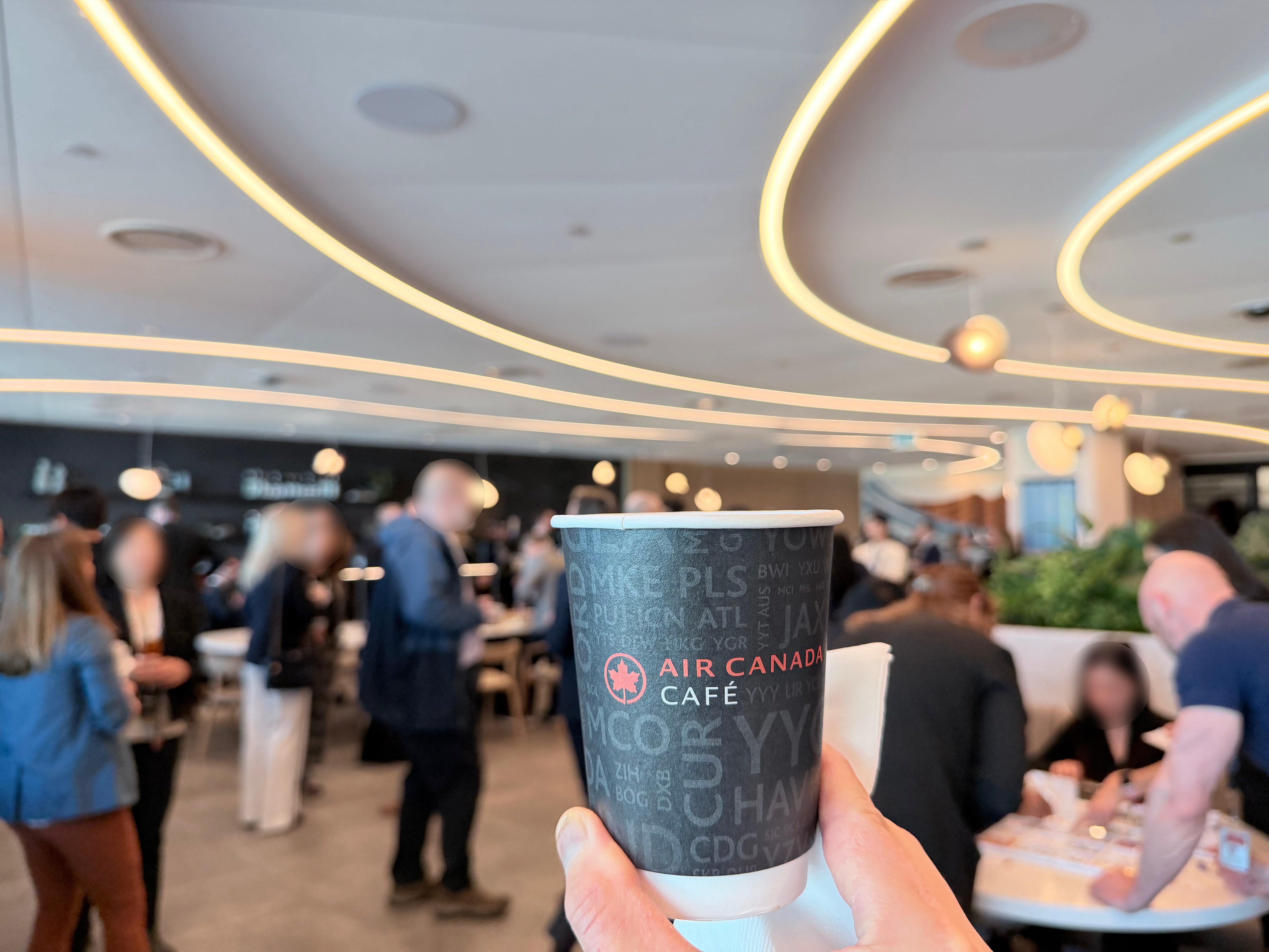 Air Canada Café branded coffee cup held up with the café interior and opening event crowd in the background