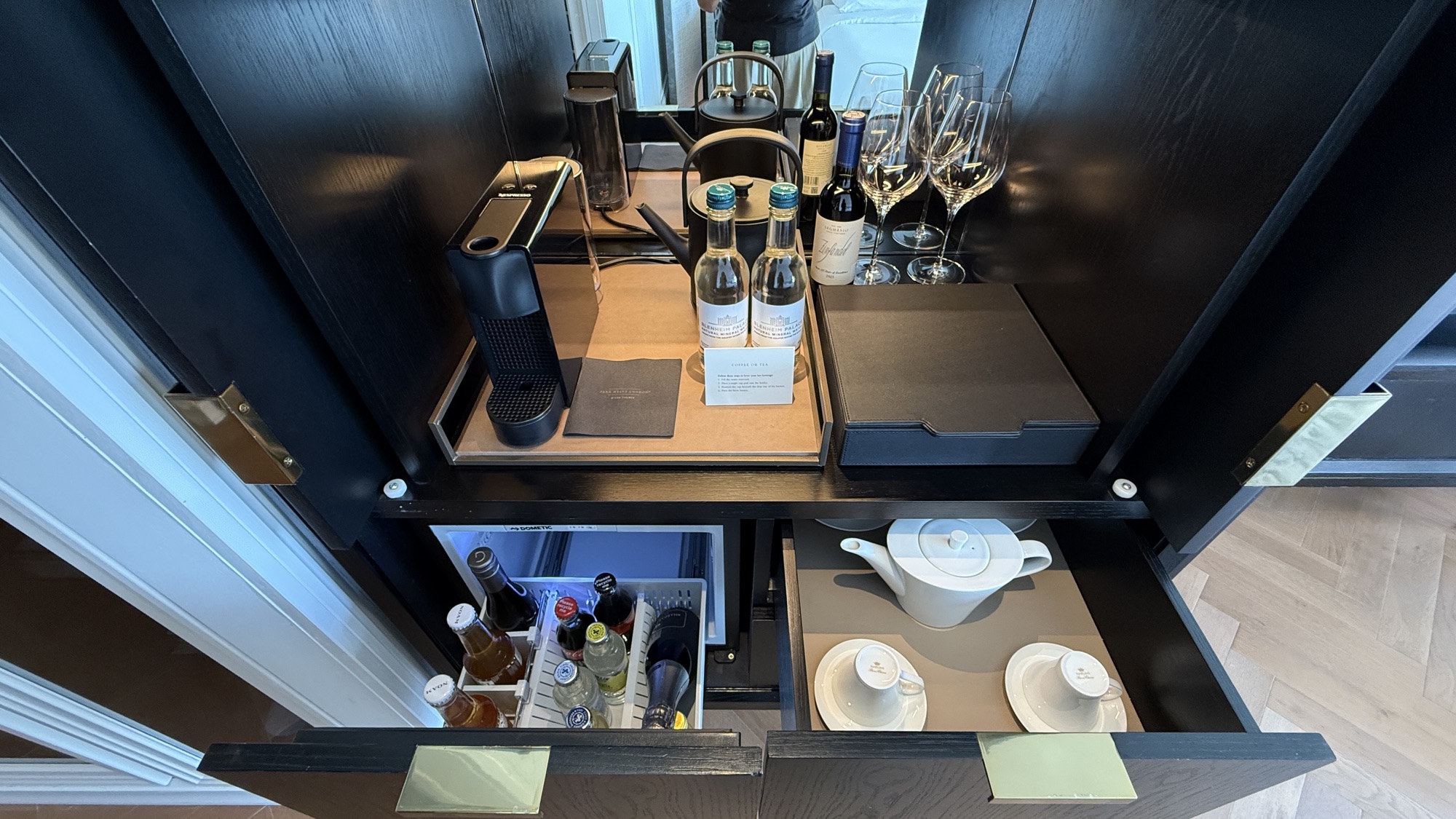 Open beverage cabinet with a Nespresso machine, kettle, Blenheim Palace mineral water, red wine and glasses on top, and a stocked Dometic minibar and teapot drawer below