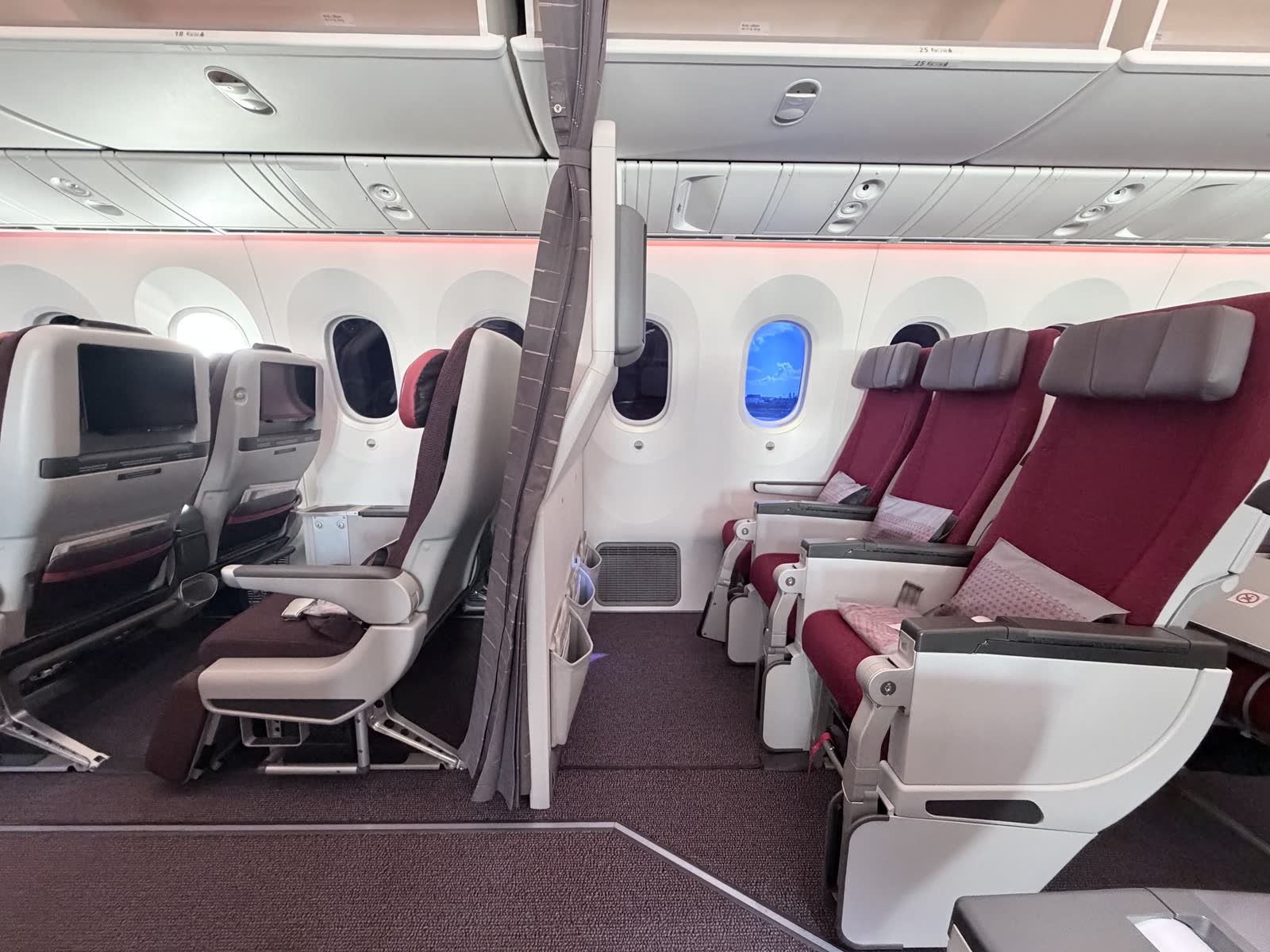 Side view of a burgundy premium economy seat and the first row of magenta economy seats, split by a partition and grey curtain