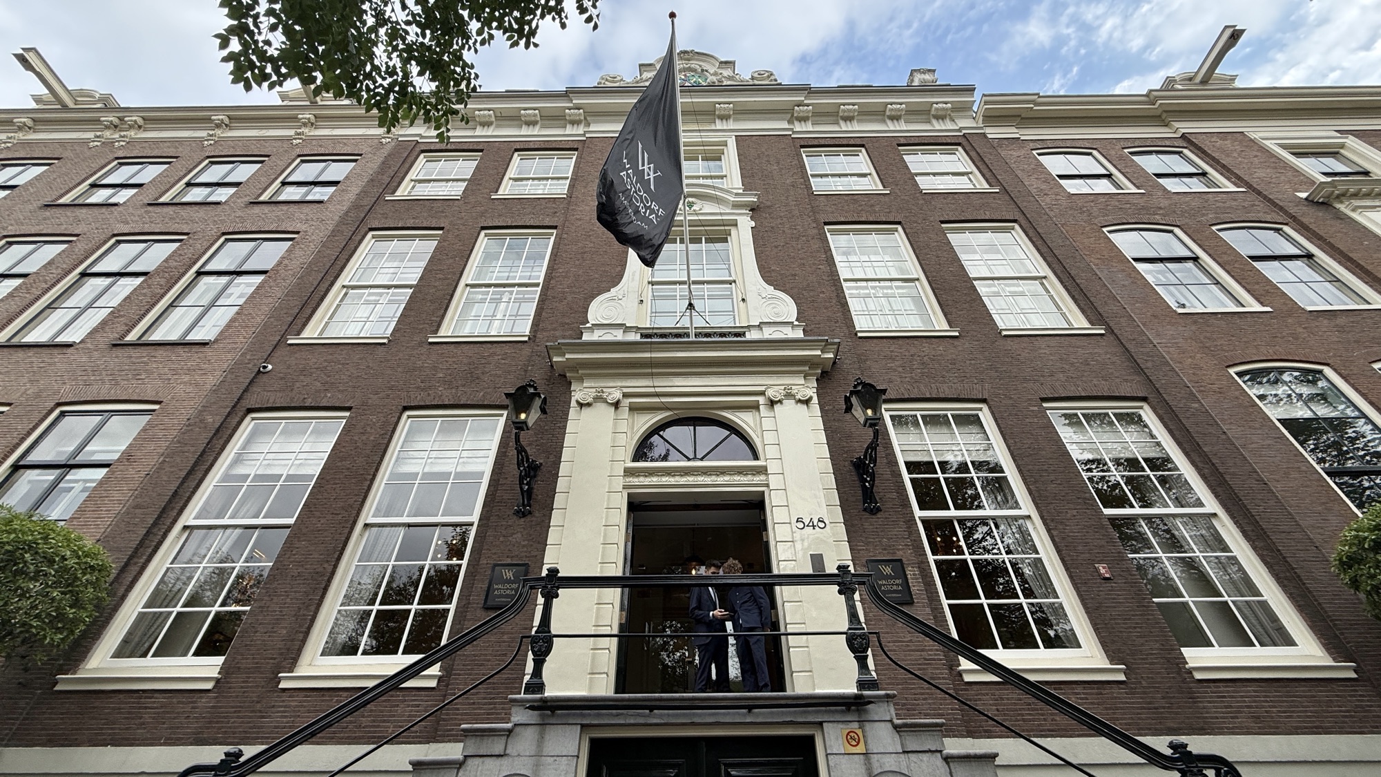 Main entrance of the Waldorf Astoria Amsterdam beneath a black flag on the brick canal-house facade